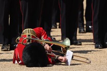 A soldier in dress uniform, holding a trombone, lies on the ground after falling.