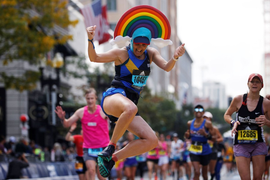 A runner wearing a rainbow hat leaps across the finish line of the Boston Marathon.
