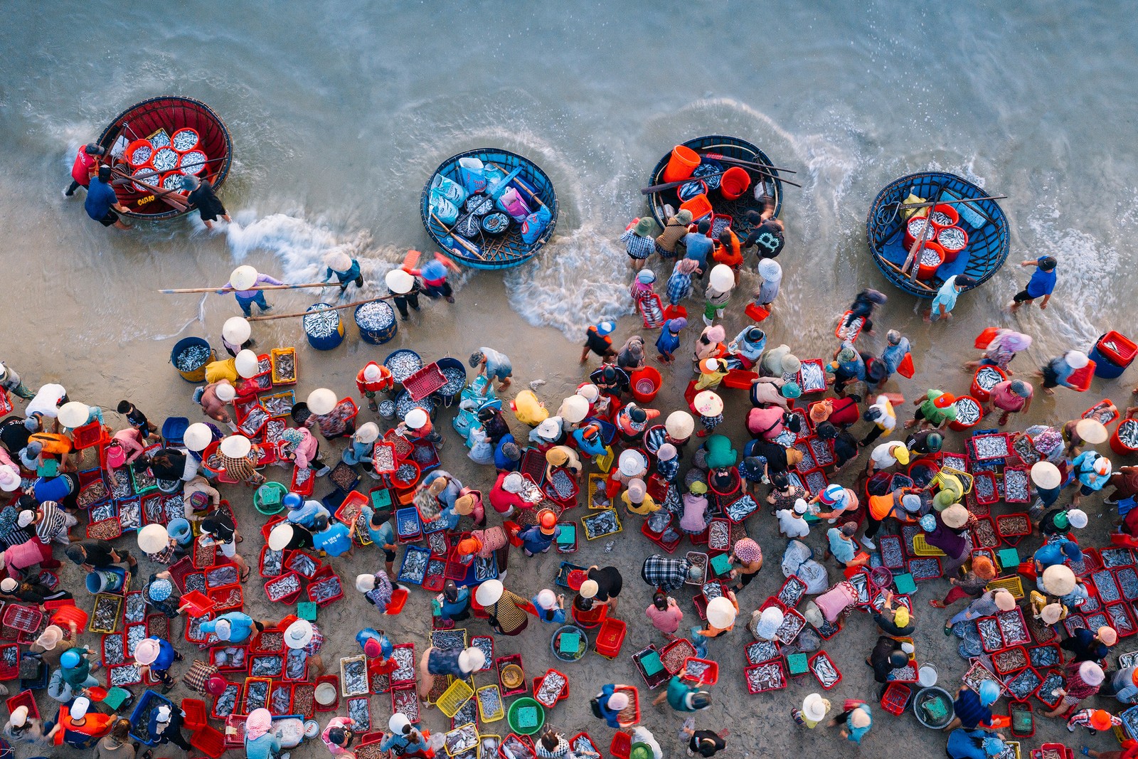 An overhead view of many people with baskets of fish standing on a shoreline.