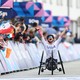 An athlete in a racing wheelchair raises their arms in celebration as they pass a crowd at a finish line.