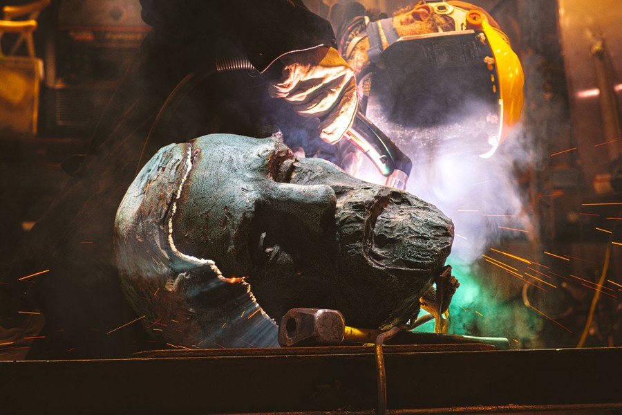 A worker uses a plasma torch to cut apart the head of a bronze statue.