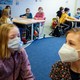 Two young girls with masks on are near the camera, and more children with masks can be seen at tables in the background