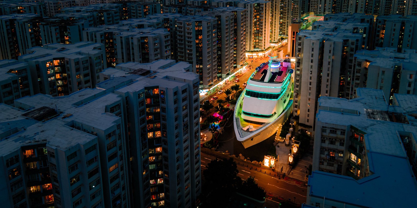 An elevated view of a large boat-shaped building sitting in a plaza among many other multi-story residential buildings.