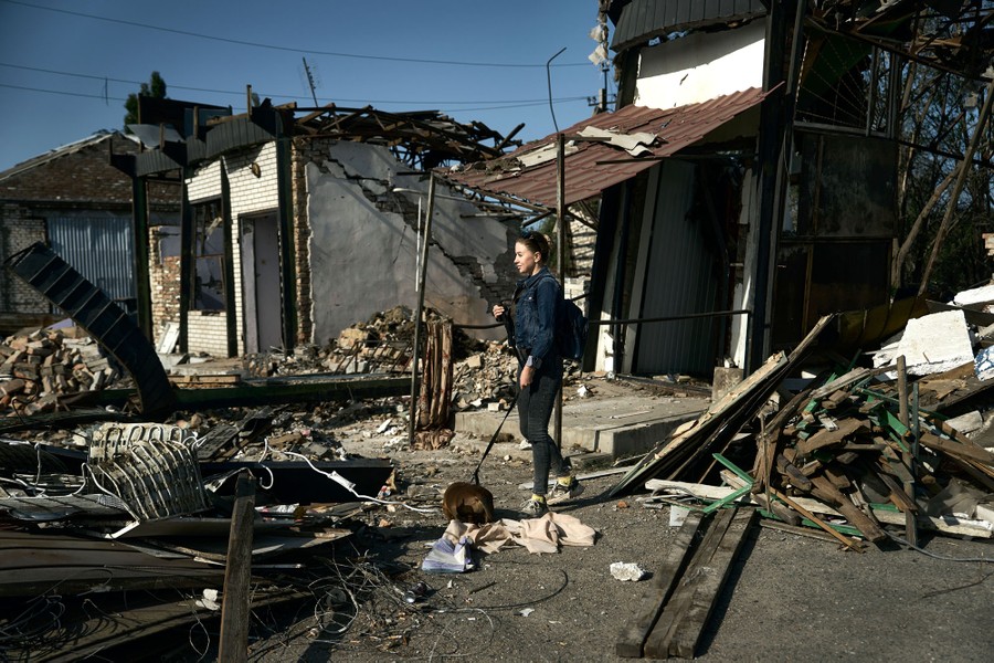 A person walks with a small dog on a leash, through the rubble of several destroyed buildings.