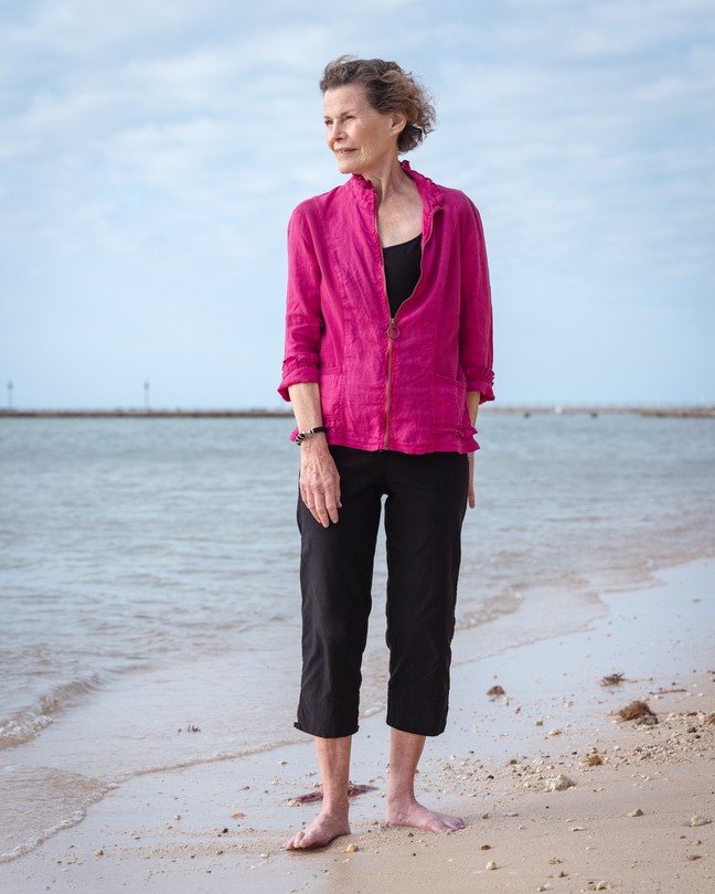 Woman in magenta top and black capri pants standing barefoot on beach with cloudy blue sky behind
