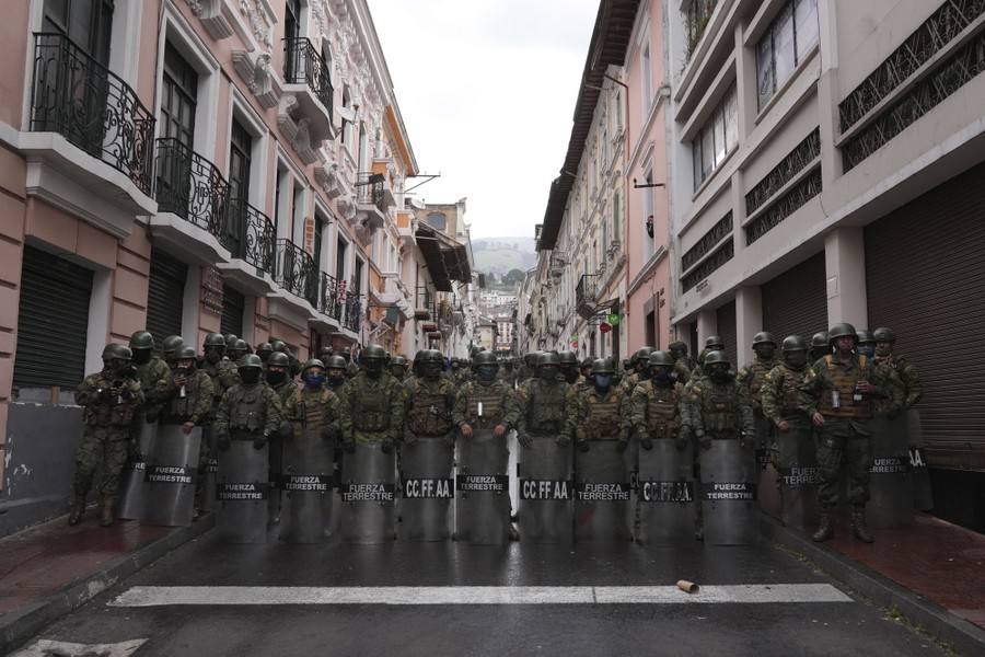 Officers in military gear stand with shields, blocking a city street.
