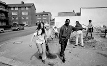 Image of two young women and one young man carrying a trash can together down a neighborhood sidewalk.