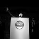 Black-and-white image of Giorgia Meloni raising her arm in the air behind a podium as she talks into the microphone, surrounded by three men clapping