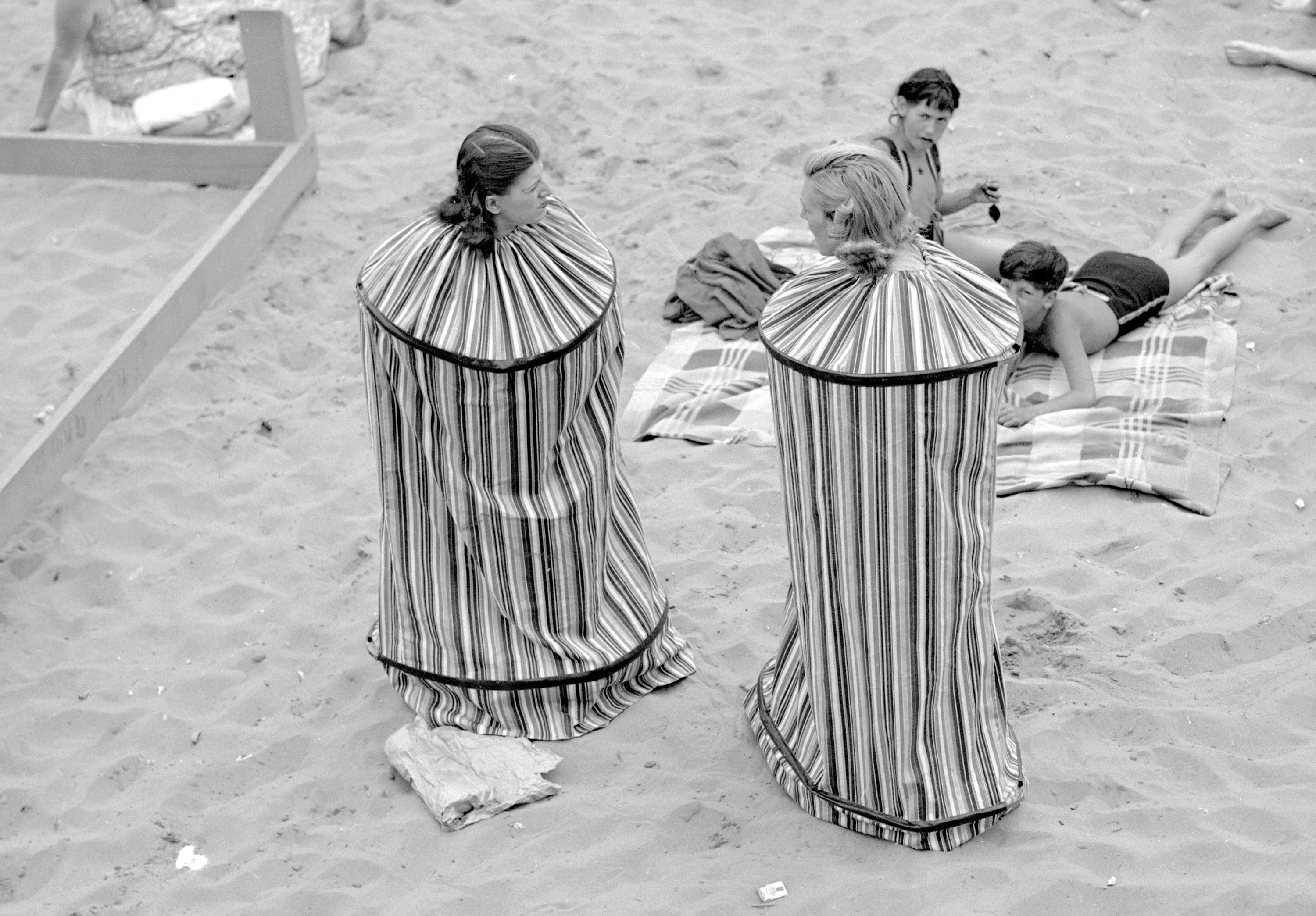 Two women stand on a beach, each wearing what looks like a small personal tent—a