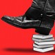 A black cowboy boot steps on a pile of books, against a red background