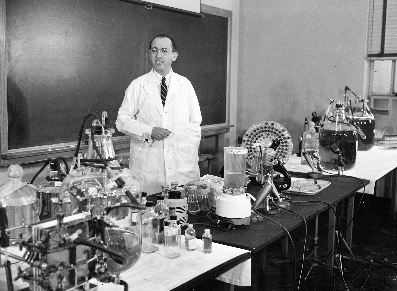 A scientist in a white lab coat stands behind tables lined with medical gear and a microphone.