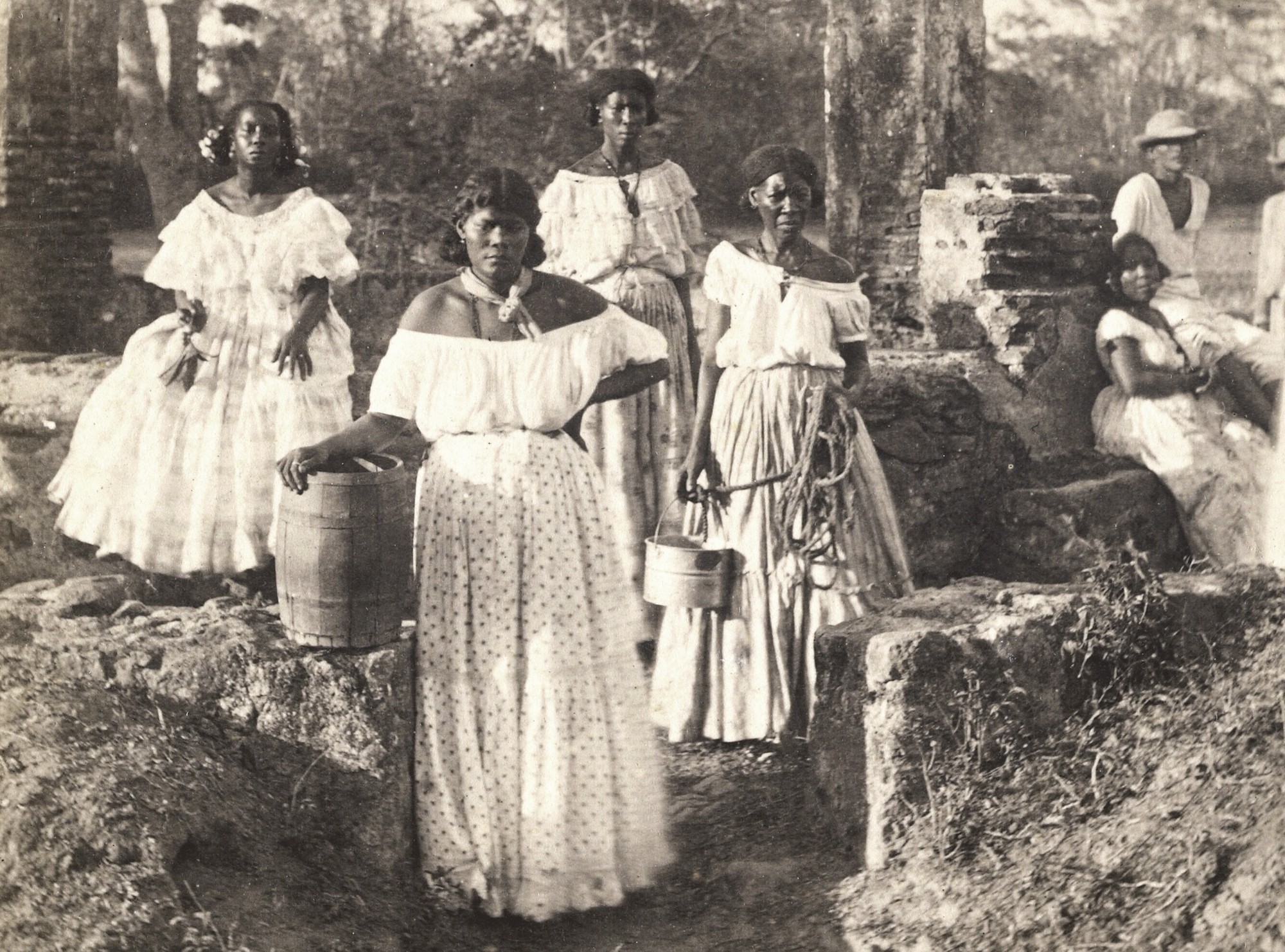 Five women and a couple of men sit and stand outside, posing in white clothing. One holds a bucket and long rope.