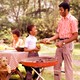 A family grills meat next to a picnic table.