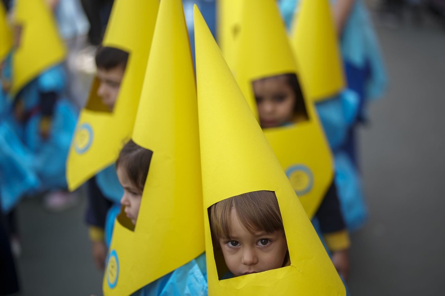 Several children march in a street wearing yellow conical hoods with a large cutout for their face.