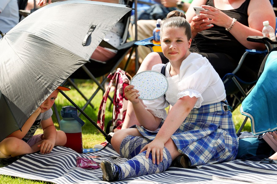 A young performer sits on a blanket with others, using a small fan to cool herself.