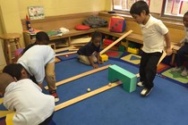 A group of pre-school students play on a blue carpet with wooden ramps and balls.