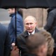 Vladimir Putin stands under an umbrella in the rain, surrounded by people facing the opposite direction.