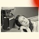 A black-and-white photo shows a high-school-aged girl reclining on her 1950s-era couch with her ear next to an old radio on a side table. Her hands are folded under her chin and she's gazing off to the right as she listens. The photo is set against a background with three spray-painted colorful circles. In the bottom left corner is a blue one, in the bottom right corner a green one and the top right corner is a red one.