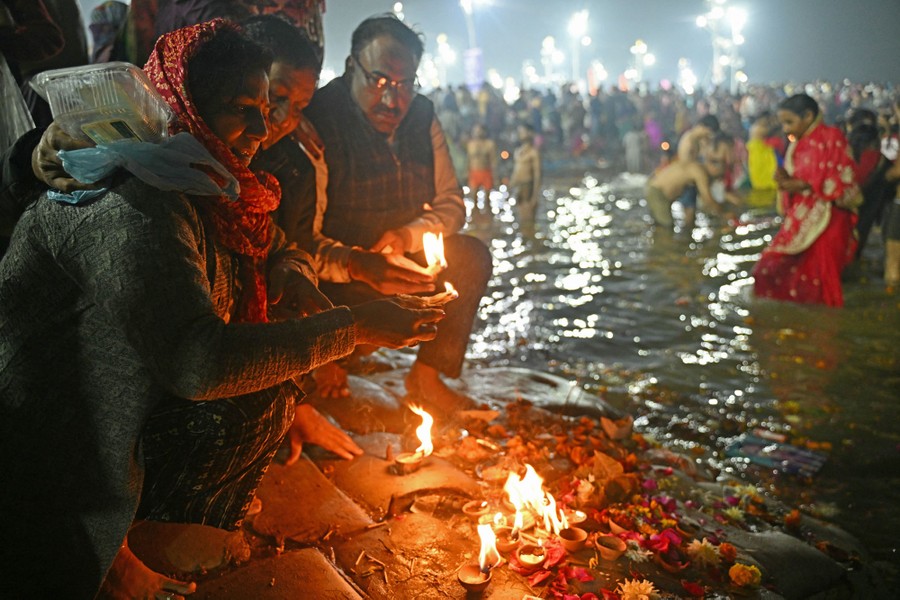 People kneel at a river's edge, lighting small lanterns, as others wade nearby.