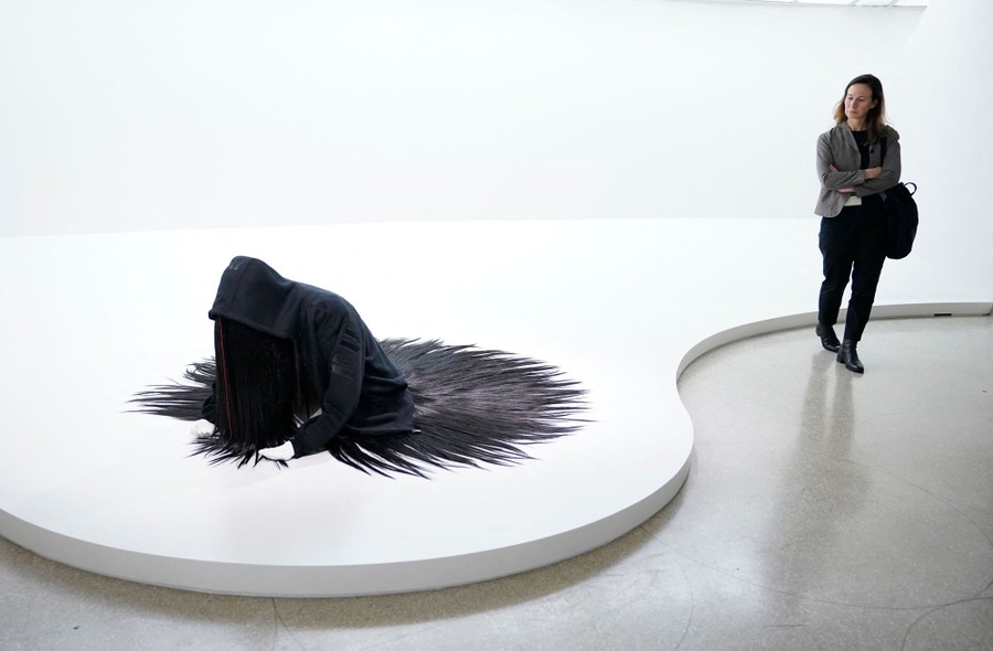 A person looks at a work of art that resembles a human being with long dark hair climbing out of the floor.