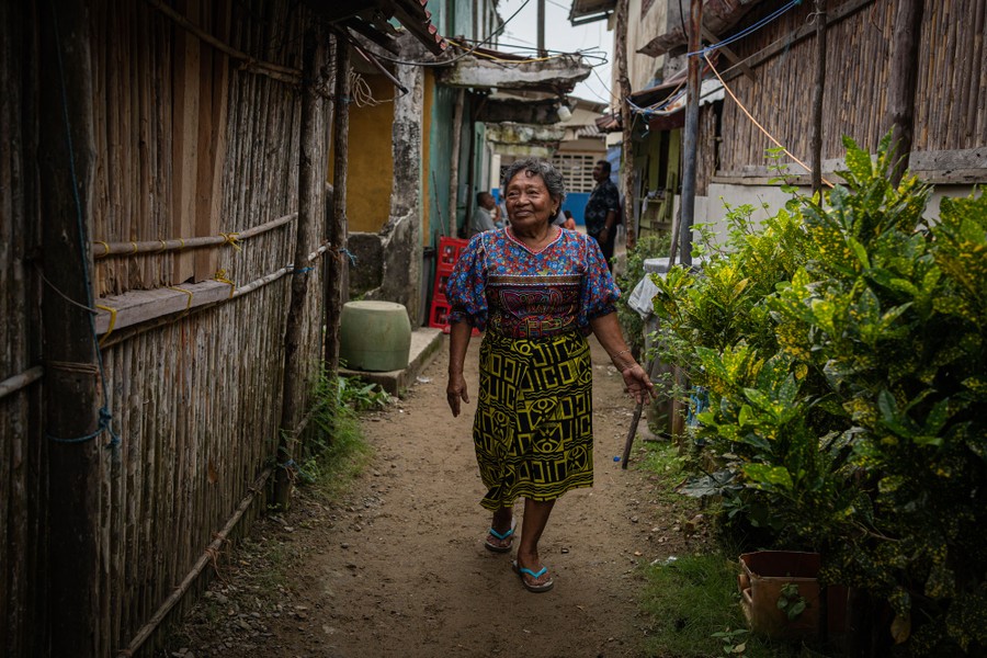 A person in a blue floral shirt and black patterned skirt walks down a dirt path between many small buildings.