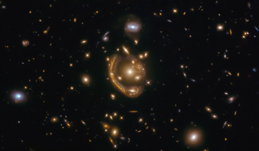 Dozens of distant galaxies are seen, with one large one at center appearing to warp and wrap itself around another bright object.