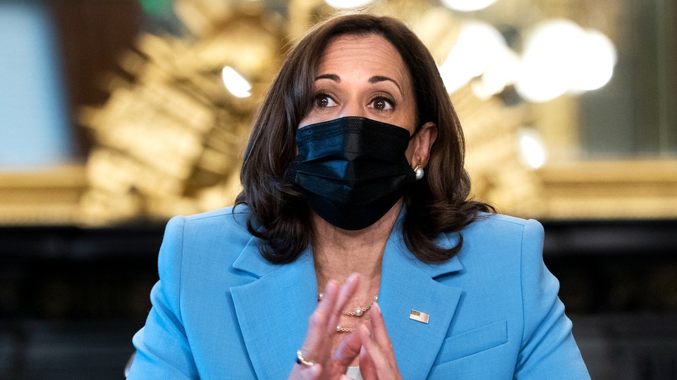 Bust photo of Kamala Harris wearing a light blue suit and a black surgical mask over a blurred background.