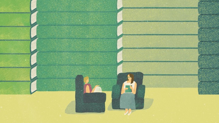 Two women sit in comfortable armchairs, in front of a wall of giant books