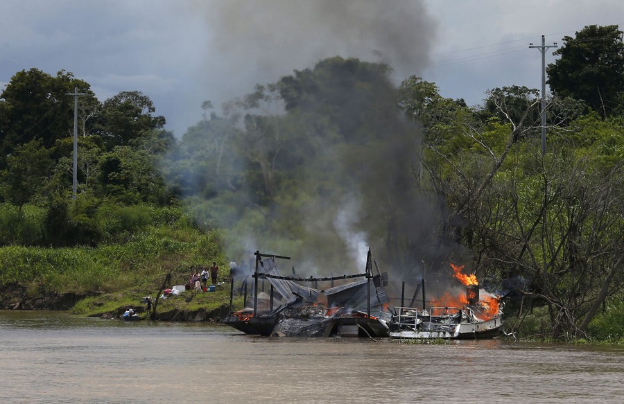 People watch a burning barge on a river shore.