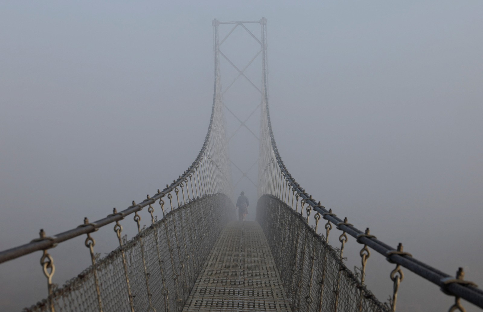 A woman walks on a suspension bridge during a foggy morning.