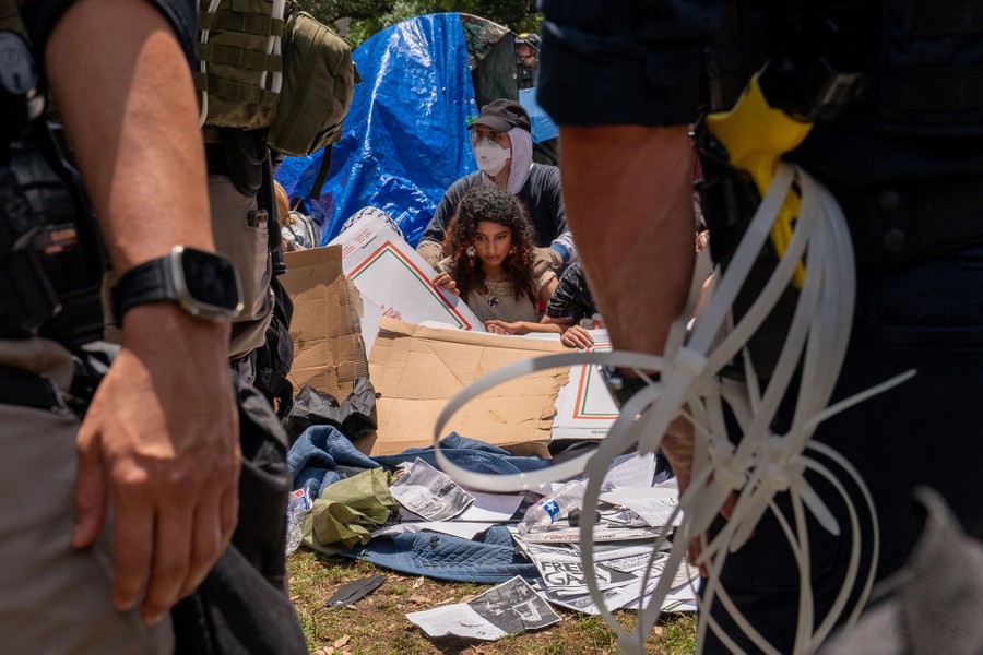 Several protesters are seen sitting on the ground, amid tents, pizza boxes, and blankets, behind police officers in riot gear.