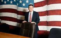 Photo of a man standing behind a desk chair against a wall painted with a large mural of the American flag