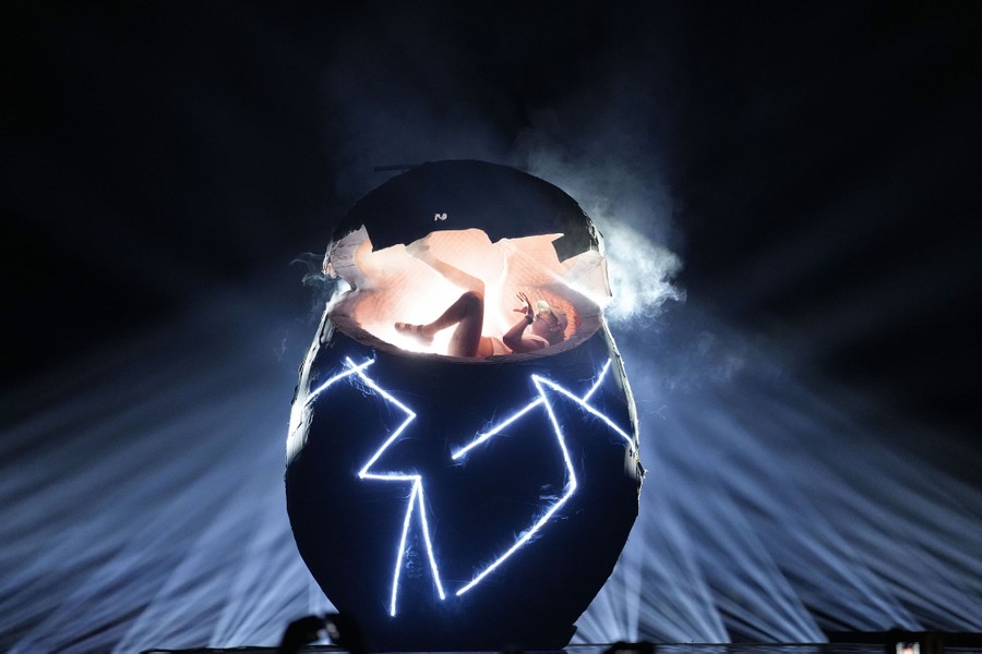 A performer sings while emerging from a large egg-shaped structure on a stage.