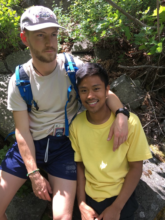 Two men sit in nature, one with his arm around the other's shoulders.