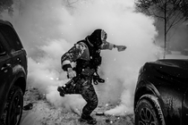 A black-and-white photo of an ICE agent between two cars and a cloud of tear gas in the background