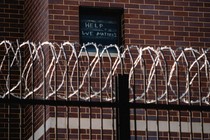 Barbed-wire in the foreground of a jail. A sign in the window reads, 'Help We Matter 2'