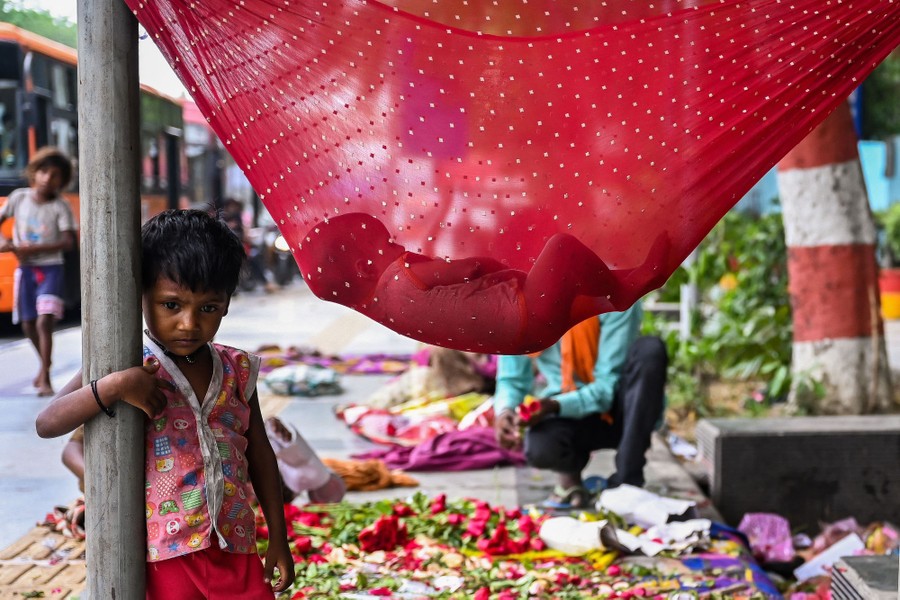 A child leans on a pole, while another smaller child sleeps in a makeshift hammock.