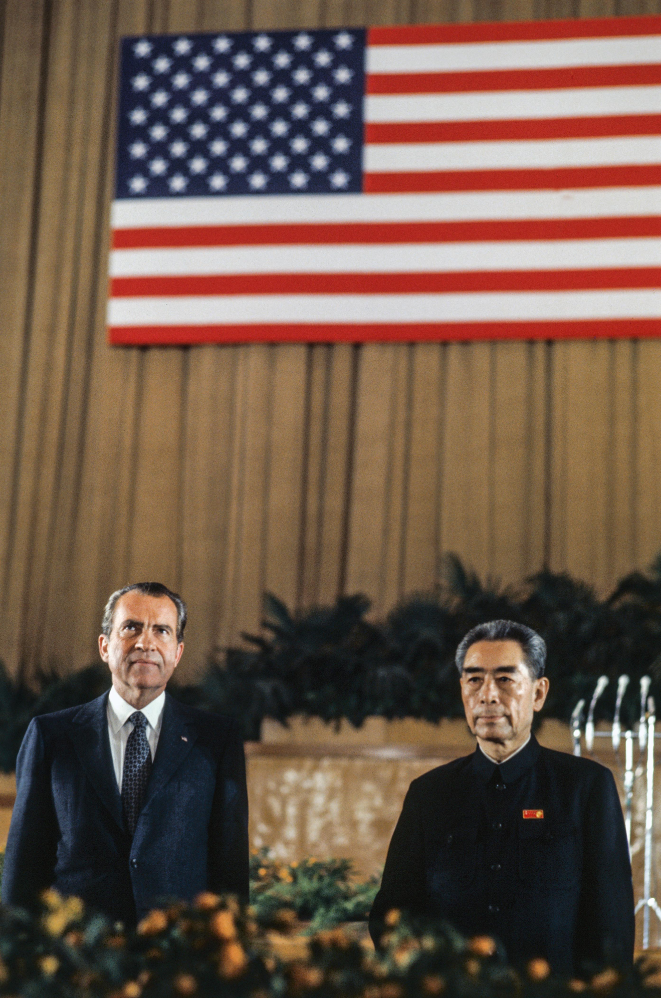 photo of Nixon and Enlai near a stage, with large curtained wall with U.S. flag displayed