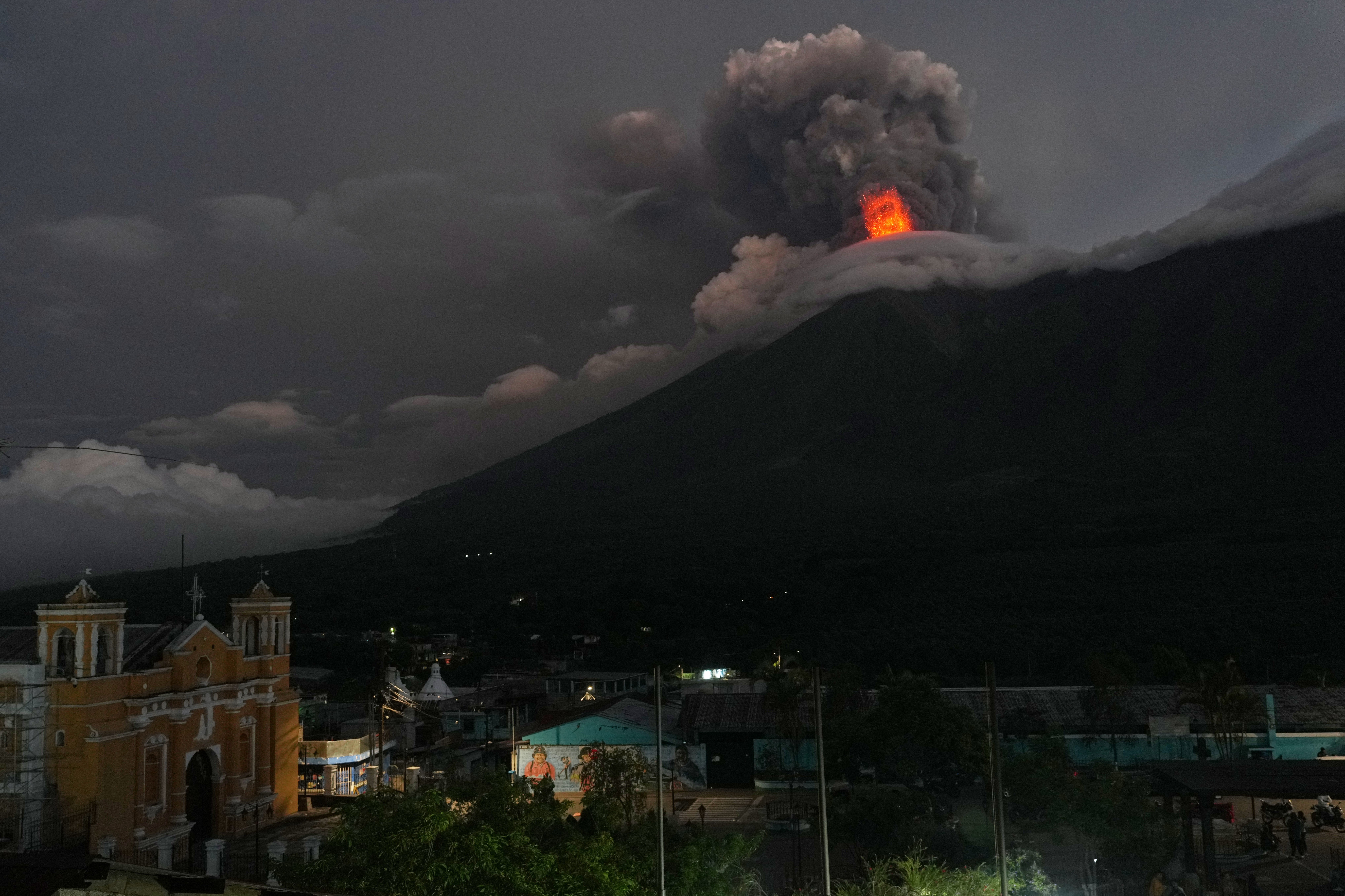 Lava and ash erupt out of the crater of a volcano, seen from a nearby village at night.