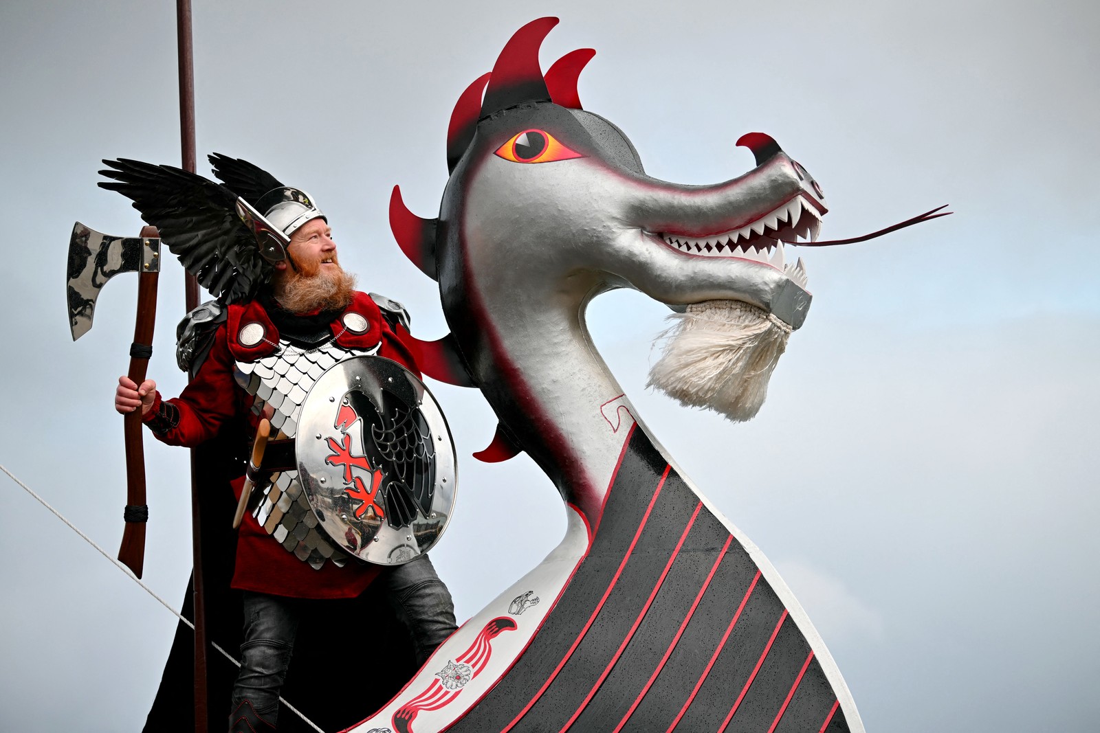 A person dressed as a Viking poses while standing beside the dragon head of a replica Viking ship.
