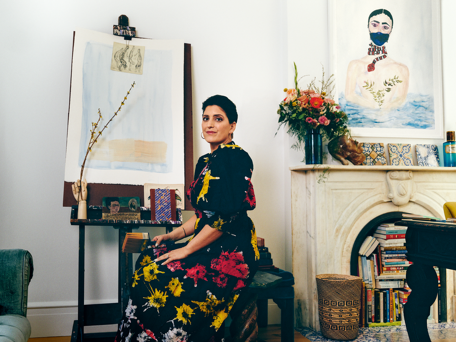 photo of dark-haired woman in red, yellow, and black dress sitting in front of easel near fireplace mantel