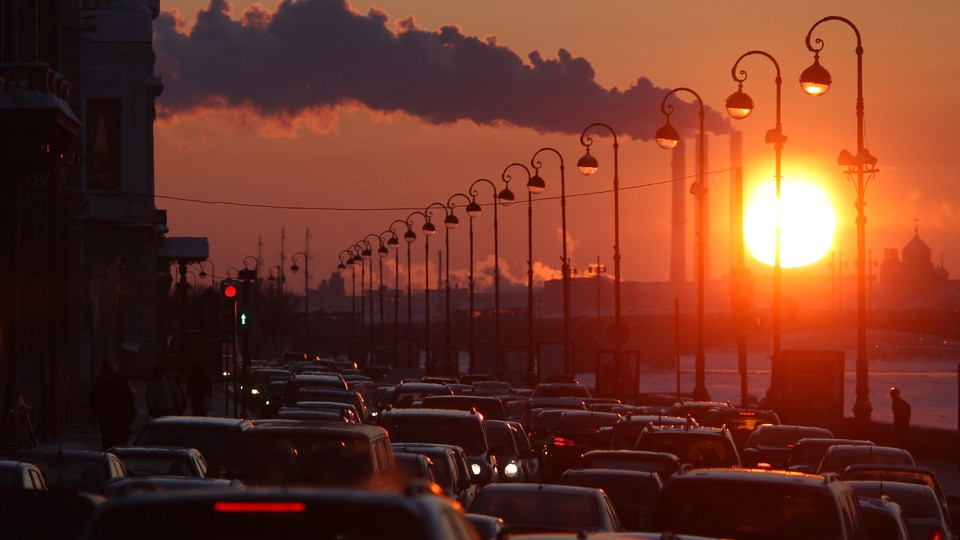 Pipes of a thermal power plant are seen during sunset, with cars stuck in a traffic jam in the foreground.