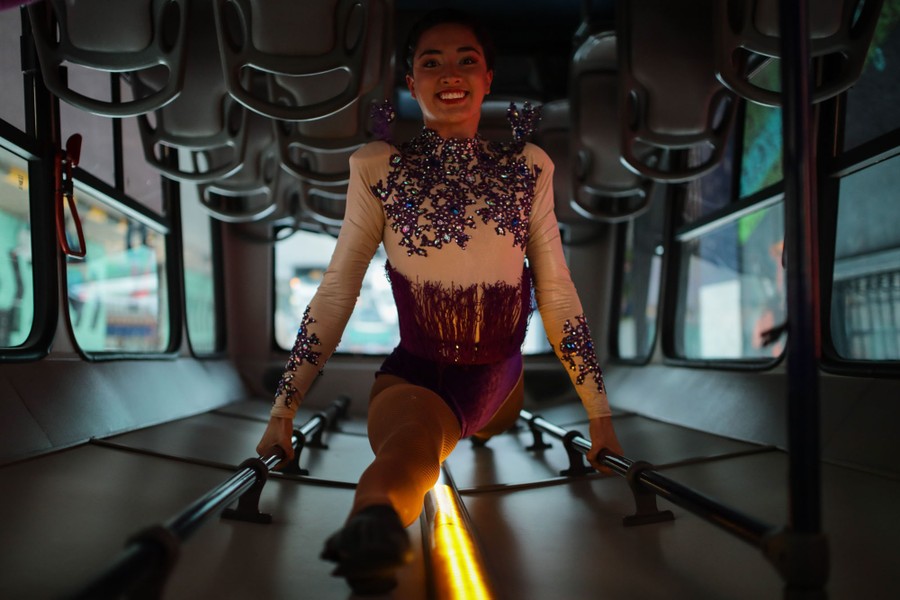 A circus performer hangs upside down from handrails inside a bus, doing the splits on the ceiling.
