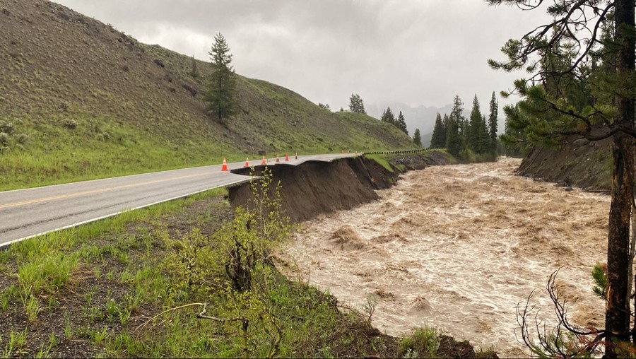 A fast-flowing river erodes its banks, undercutting and damaging part of a two-lane road.