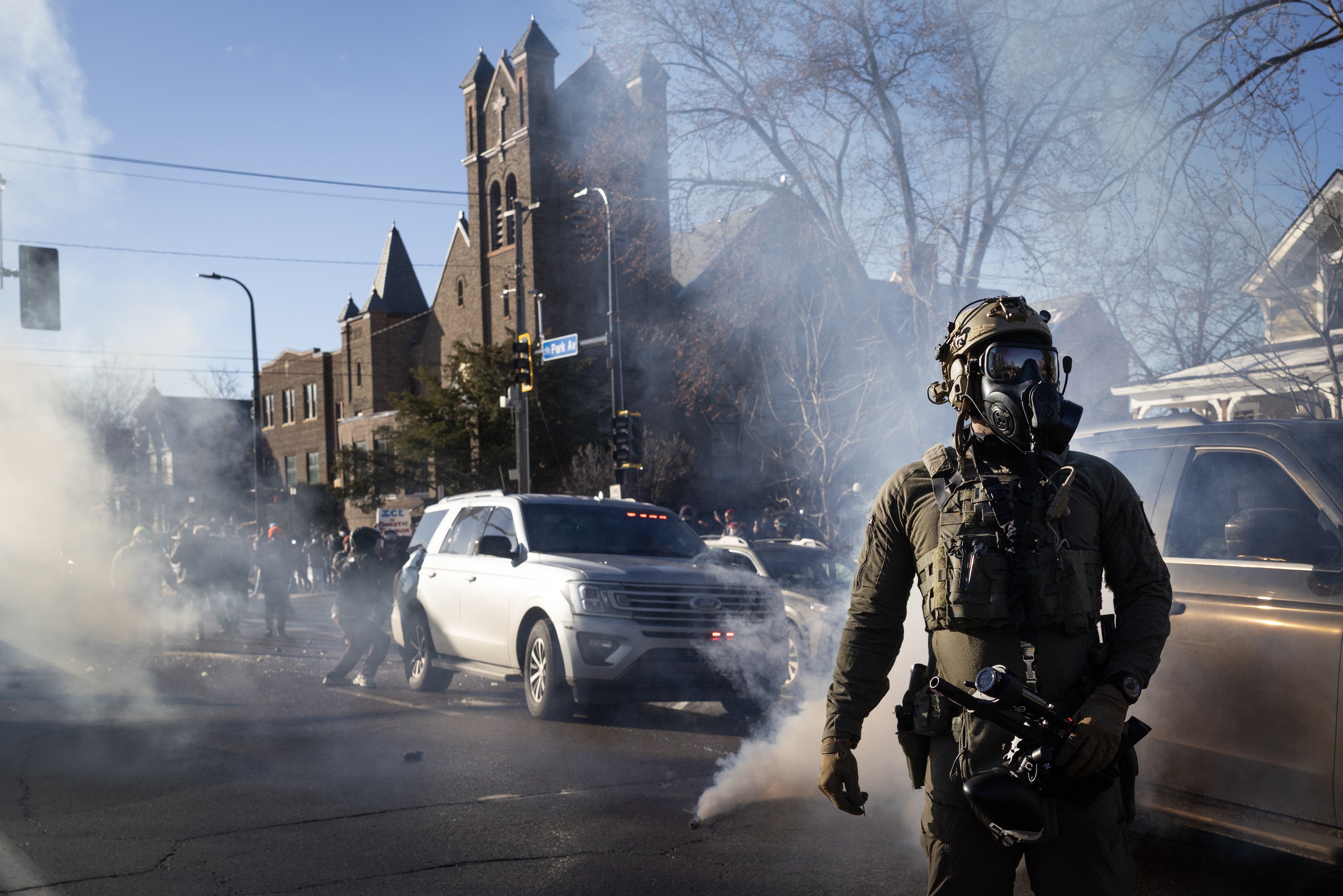 A masked federal agent stands in the foreground as tear gas fills an intersection in the background, in front of a church in a residential neighborhood.