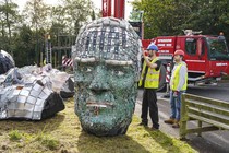 Two workers move a large sculpture—a head made of recycled material.