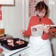 A woman reads a cookbook next to meat cooking on the stove.
