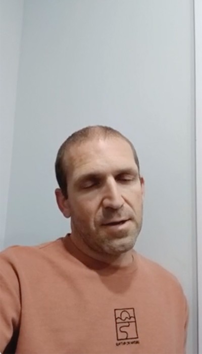 phone video screenshot of man with eyes downcast in room with gray walls