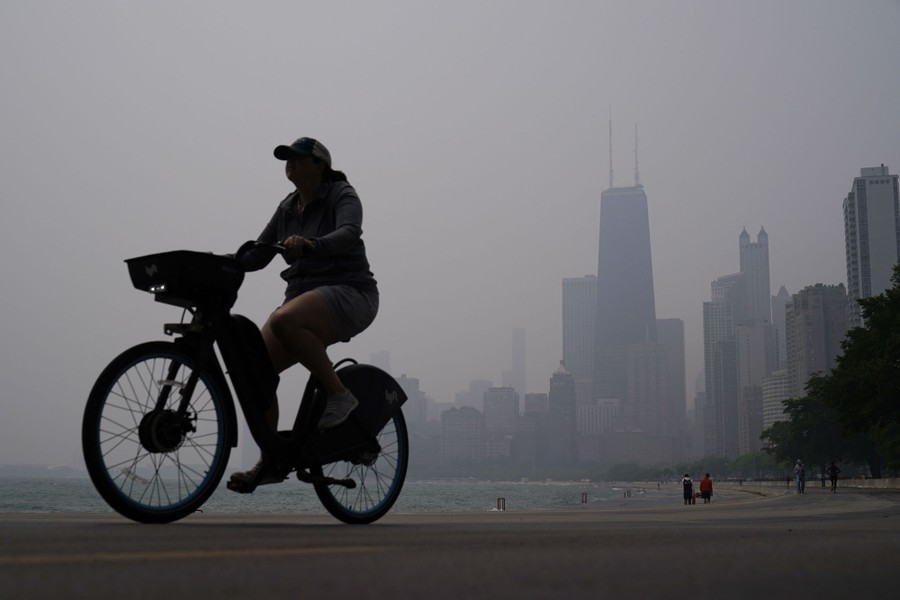 A person rides a bicycle along a shore in front of Chicago skyscrapers and a hazy sky.