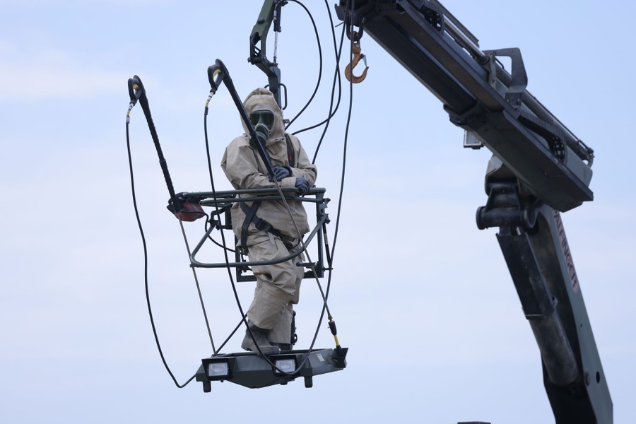 A person wearing full-body protective gear hangs in a basket beneath a crane arm.